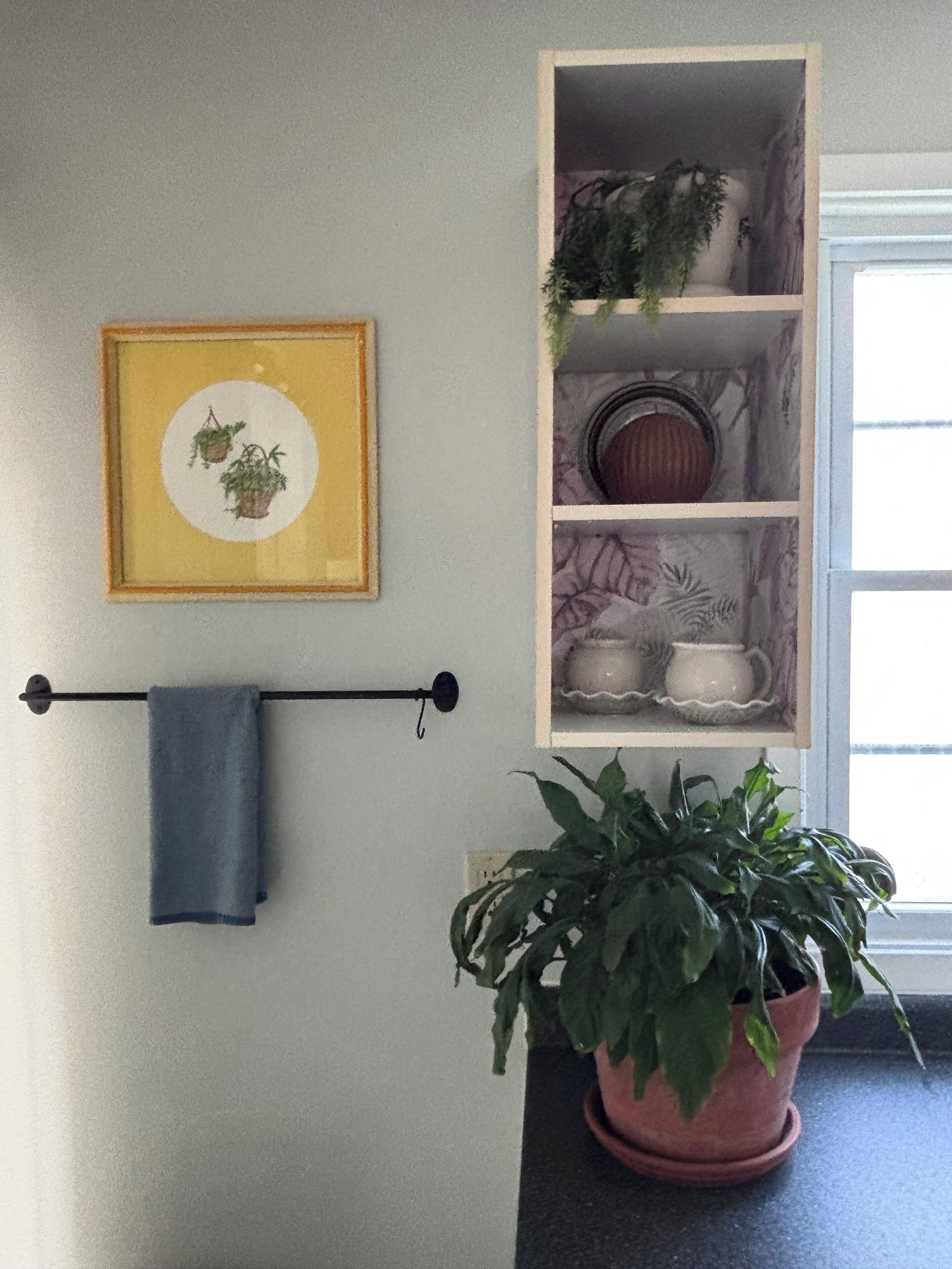 Un baño con toallero, un cuadro enmarco, repisas con objetos decorativos y una planta en maceta.