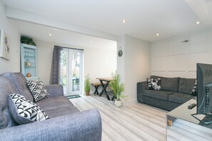 Una sala de estar moderna con un sofá gris, un sofá gris más pequeño, una mesa de centro de cristal y un televisor. Hay una mesa de comedor de madera con sillas, una planta en maceta y un ventanal corredero que da a un área exterior.