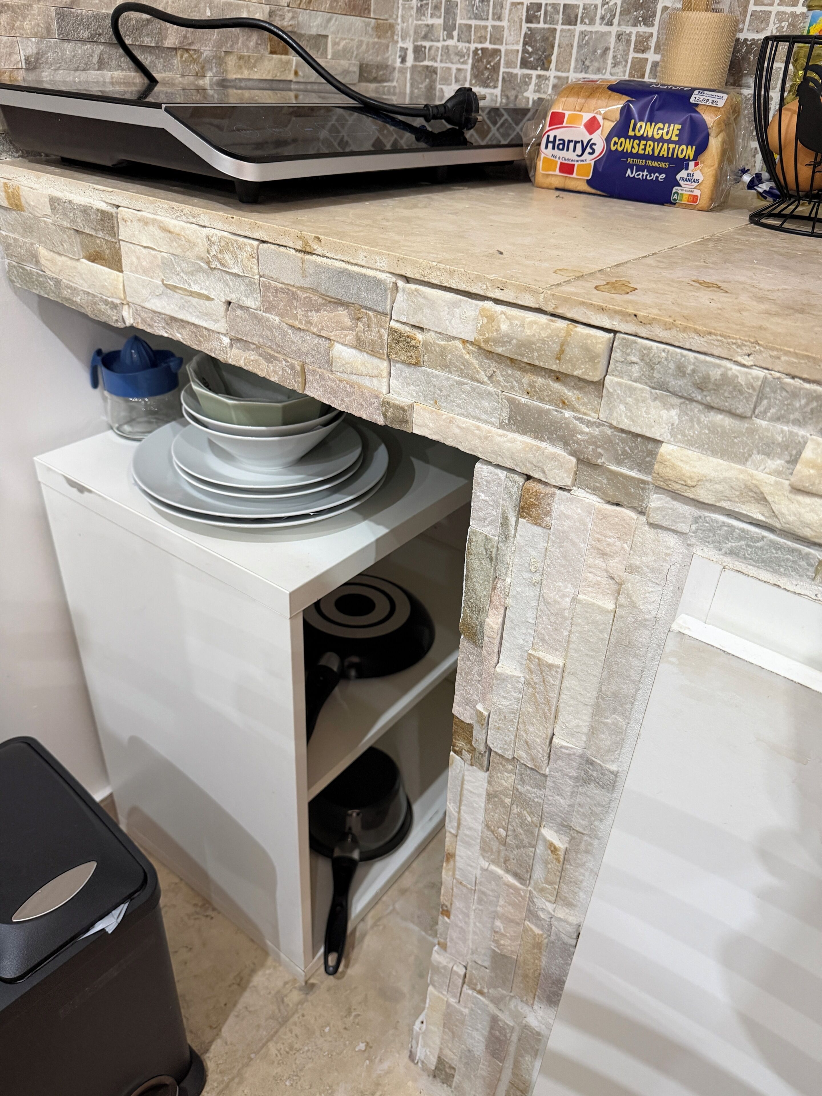 A kitchen with a stone backsplash, a bread loaf, and a trash bin.