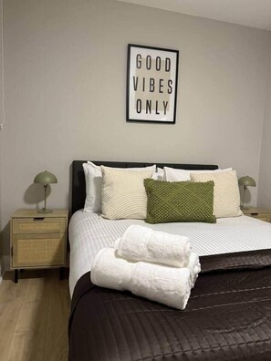 Een net opgemaakt bed met een donker hoofdbord, een groen kussen en een witte handdoek ernaast. Op de muur boven het bed hangt een lijst met de tekst 'GOOD VIBES ONLY'.