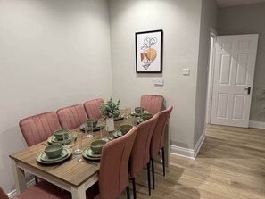 Uma sala de jantar com mesa de madeira posta para seis pessoas, cadeiras cor-de-rosa e uma obra de arte emoldurada na parede.