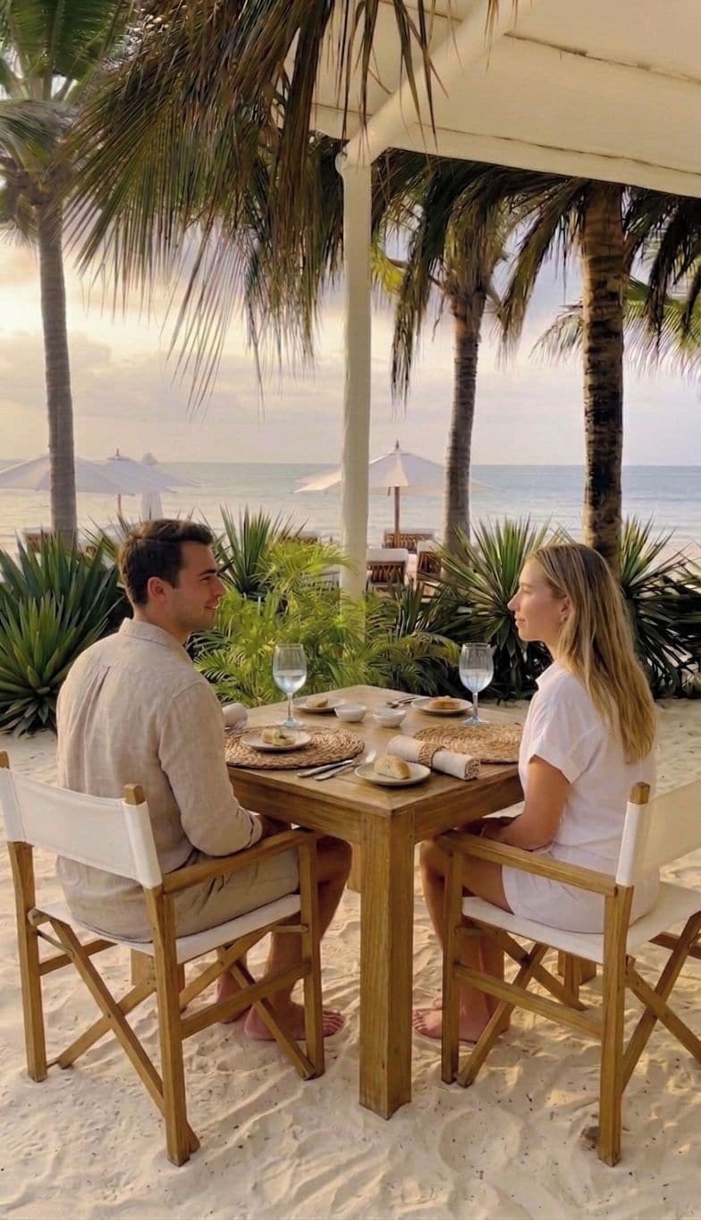 Um casal jantando em uma mesa de madeira com vista para o oceano e palmeiras.