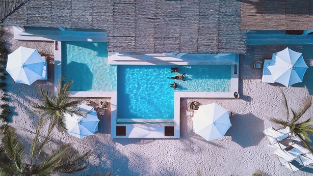 An aerial view of a swimming pool surrounded by white umbrellas and lounge chairs, with palm trees and a wooden deck.