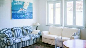 A room with a striped sofa, a plain sofa, a clear chair, a table, a lamp, and a painting of a sailboat. - Hostdomus - Brezza Marina House (Moglio)