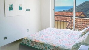 A bedroom with a bed, a window with a view of a coastal landscape, and two framed pictures on the wall. - Hostdomus - Brezza Marina House (Moglio)