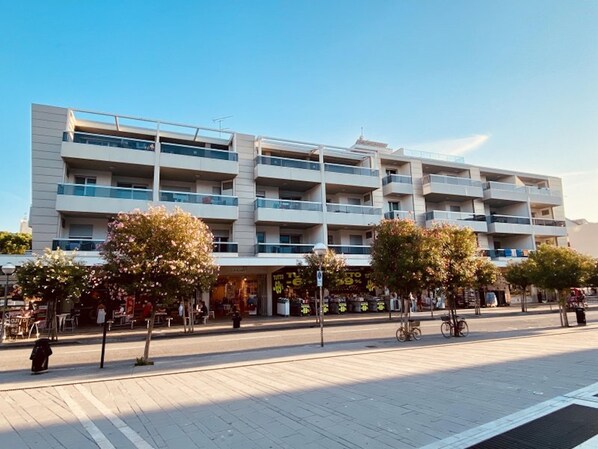 Un edificio de varios pisos con balcones, una zona peatonal con árboles y comercios.