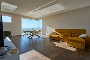 Ein modernes Wohnzimmer mit einer Couch, einem Esstisch mit Stühlen, einem Fernseher und einem großen Fenster mit Aussicht.
