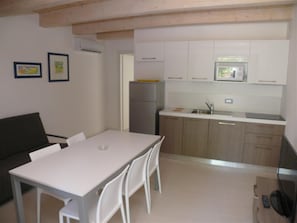 Eine kleine Küche mit weißen Schränken, einem Kühlschrank und einer Mikrowelle. Es gibt einen Esstisch mit Stühlen.