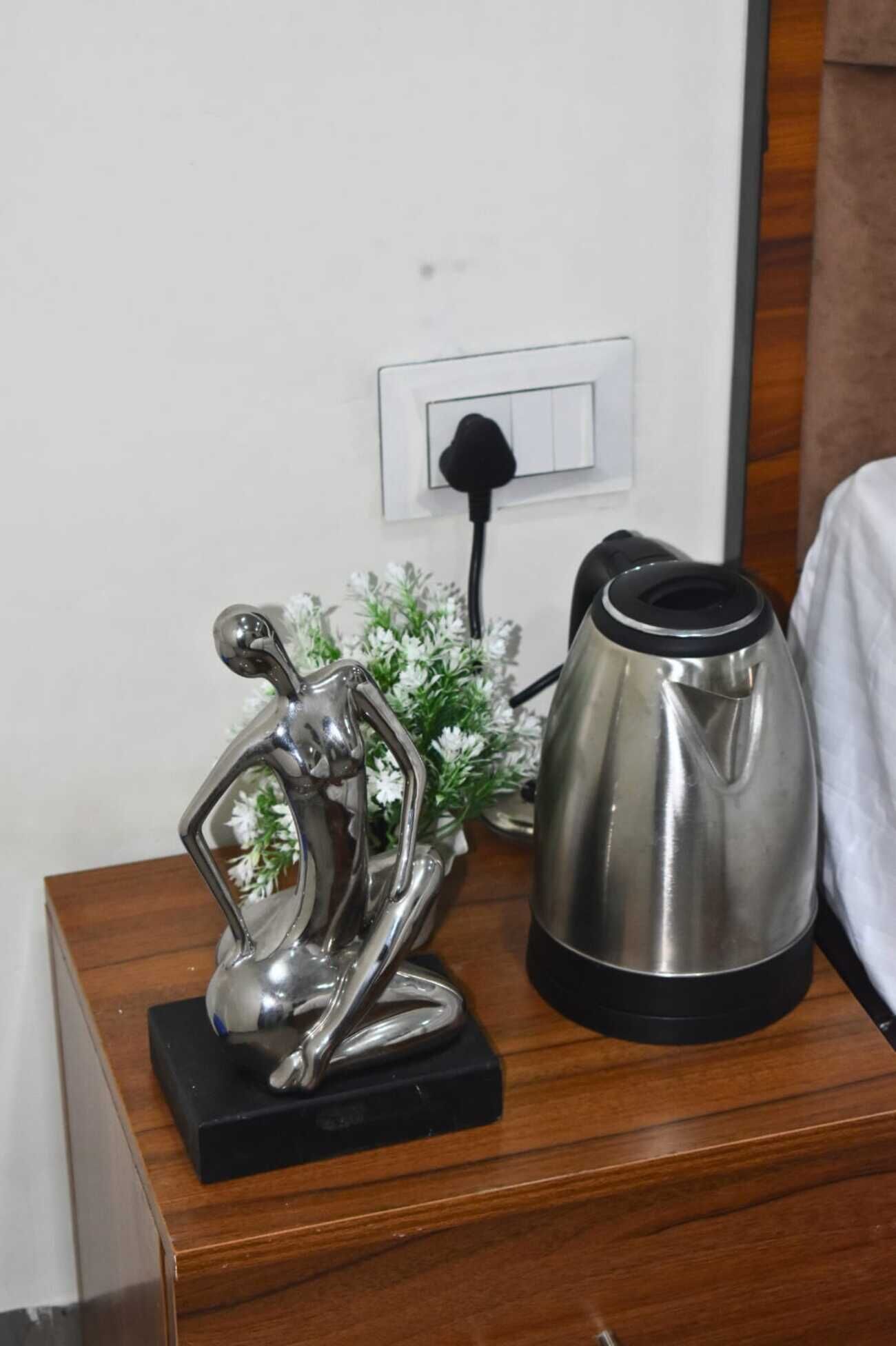 A metallic coffee pot on a wooden nightstand with a power outlet and a switch in the background.