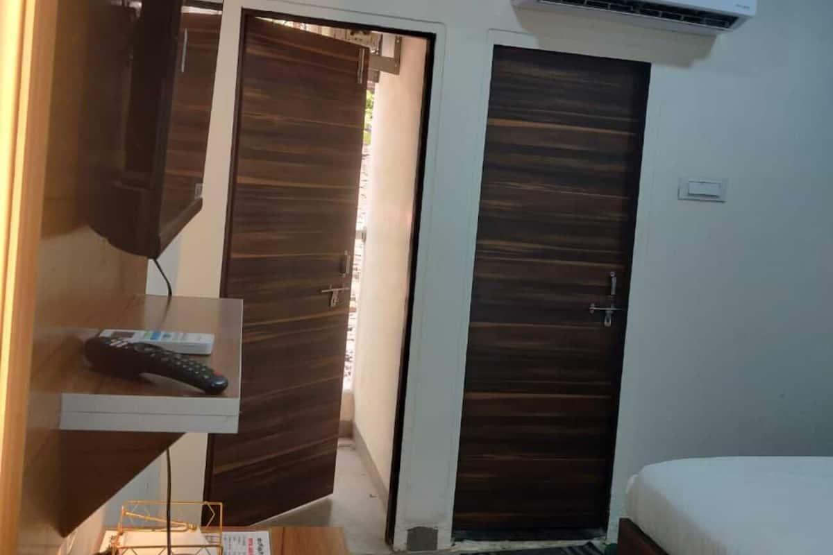 Habitación de hotel con cama, televisor montado en la pared, equipo de aire acondicionado, puerta de madera y una pequeña mesa con control remoto y vaso.
