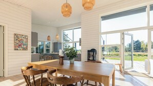 Uma cozinha espaçosa com uma ilha grande, cadeiras de madeira, luminárias pendentes e amplas janelas que oferecem vista para o exterior.