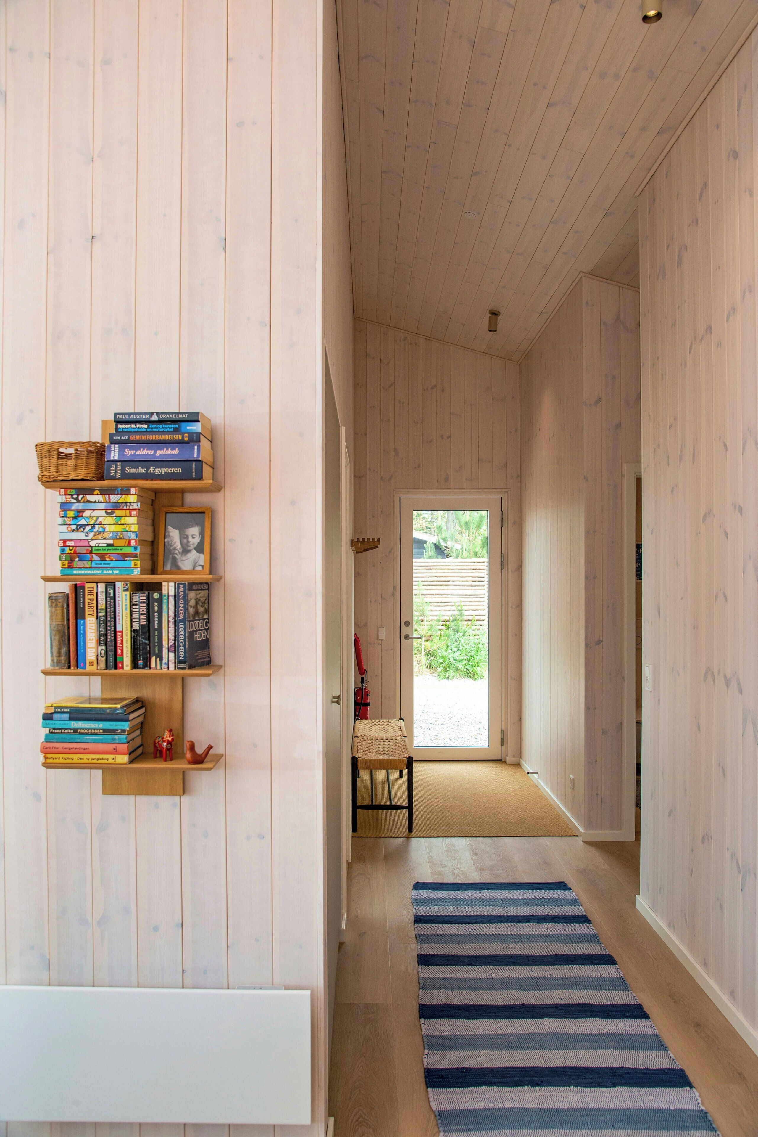 A hallway with wooden walls, a bookshelf, a blue striped rug, and a door leading to an outdoor area.