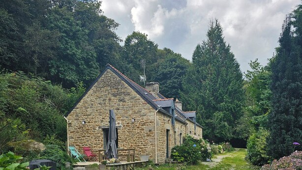 A stone building with a sloped roof, surrounded by lush greenery and a wooden gate. - Cottage in wooded heart of Brittany (Ploërdut)