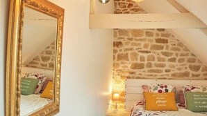 A bedroom with a sloped ceiling, a large mirror, a bed with floral bedding, and a stone wall. - Cottage in wooded heart of Brittany (Ploërdut)