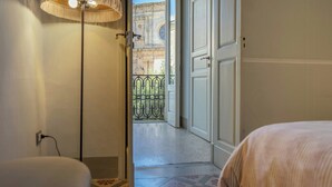 Een hotelkamer met een bed, een lamp, een deur naar een balkon met ijzeren hekwerk en uitzicht op een gebouw met versierde architectuur.