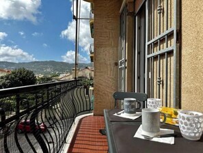 Een balkon met uitzicht op een stad en heuvels, een metalen hekwerk, een tafel met mokken en een stoel.