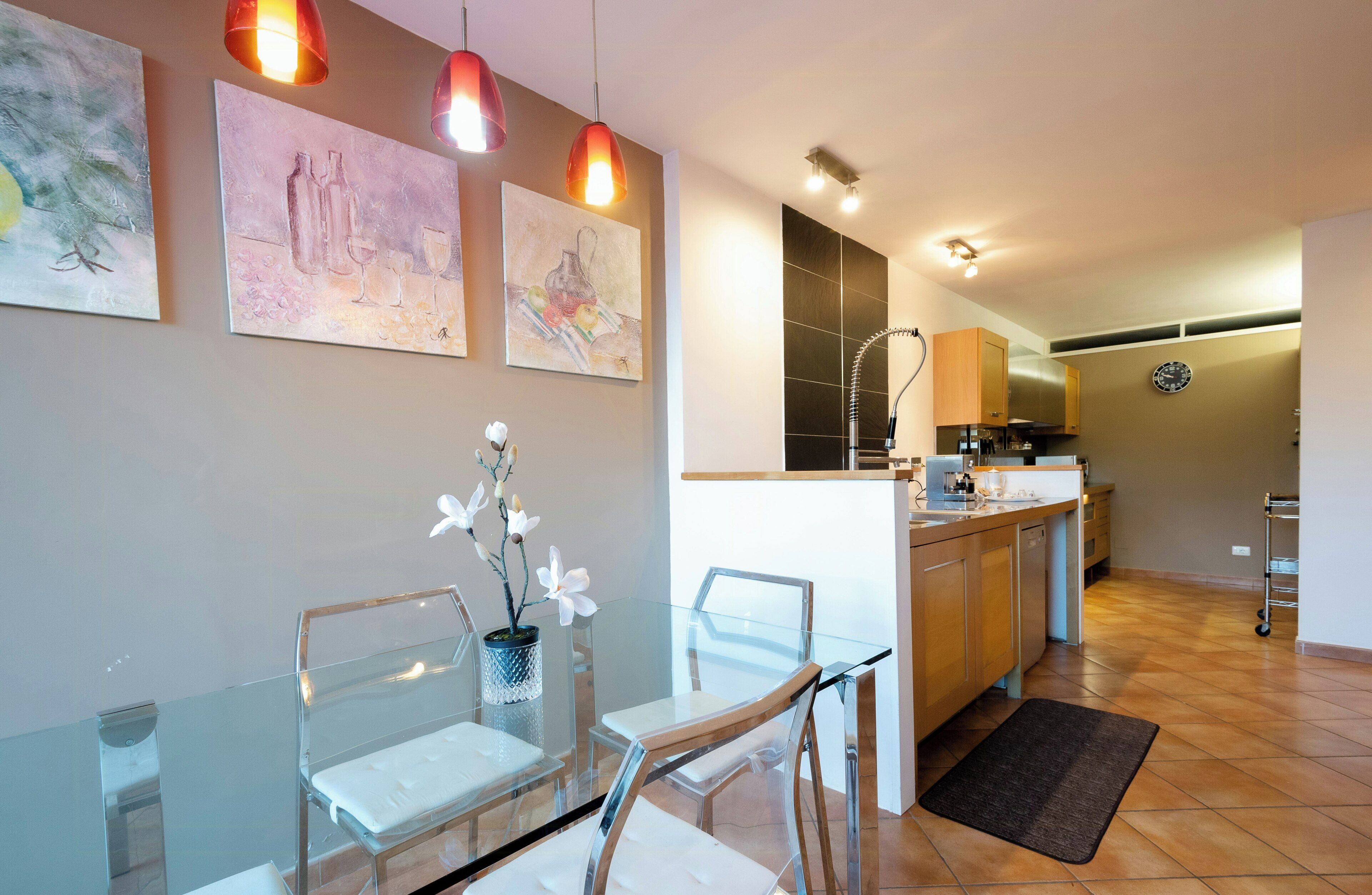 A modern kitchen with a dining area, featuring a glass table, chairs, and artwork on the walls.