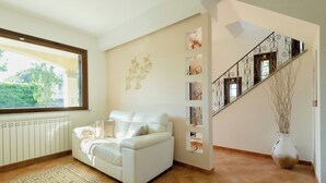 A living room with a white sofa, a patterned rug, a radiator, a window, a staircase with decorative railings, and a vase with branches. - Villa with pool at Lake Bracciano (Bracciano)