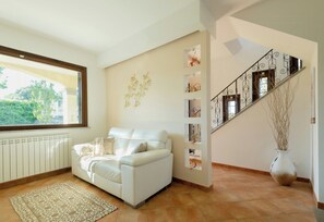 A living room with a white sofa, a patterned rug, a radiator, a window, a staircase with decorative railings, and a vase with branches.
