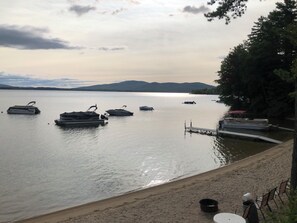 Vista de un lago con barcos amarrados, una playa arenosa y una cadena montañosa distante.