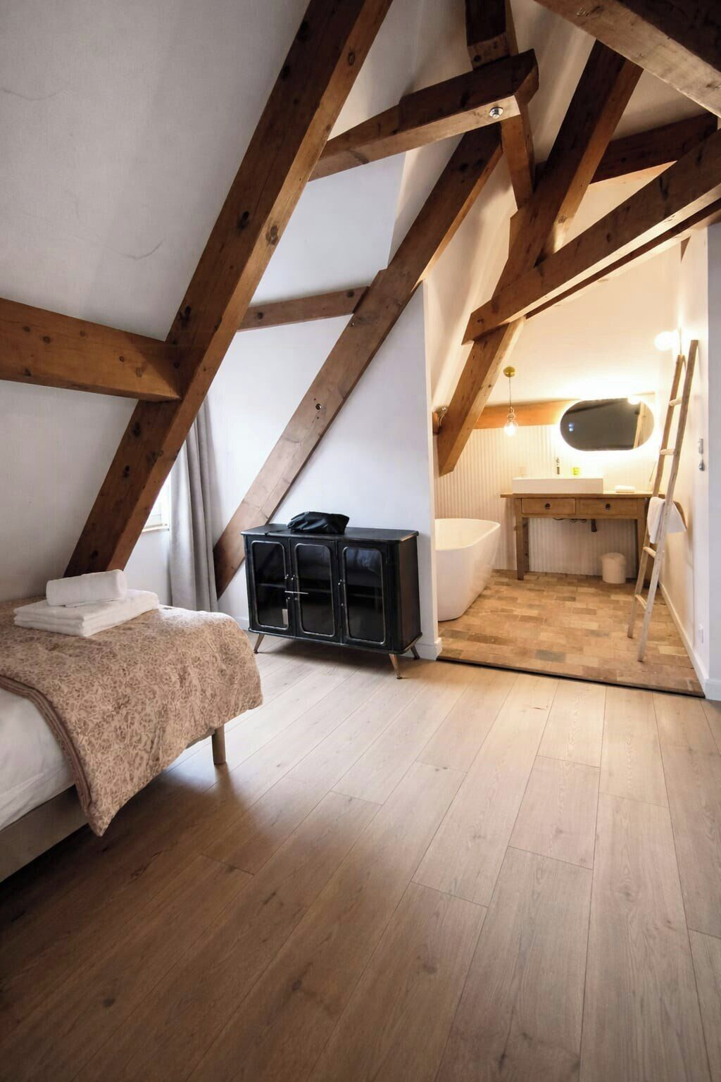 Une chambre à coucher aménagée en mezzanine avec des poutres apparentes, un lit recouvert d’une couverture à motifs, une armoire noire, une salle de bain avec un miroir et un plancher en bois.