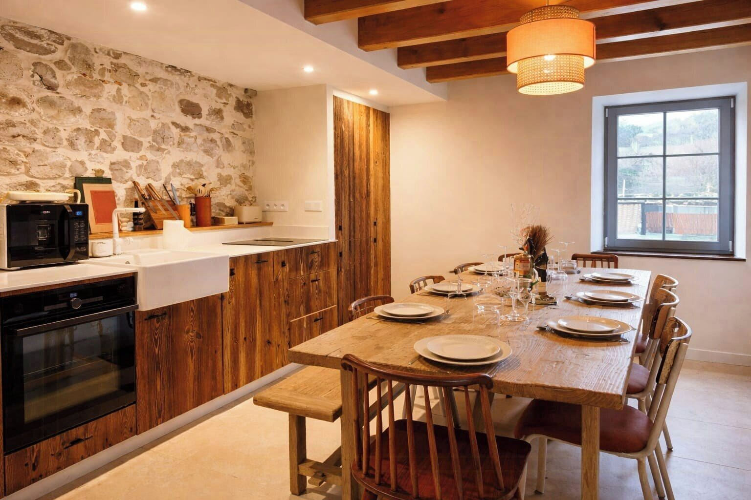 A kitchen with a stone wall, wooden cabinets, a dining table set for six, and a window with a view.