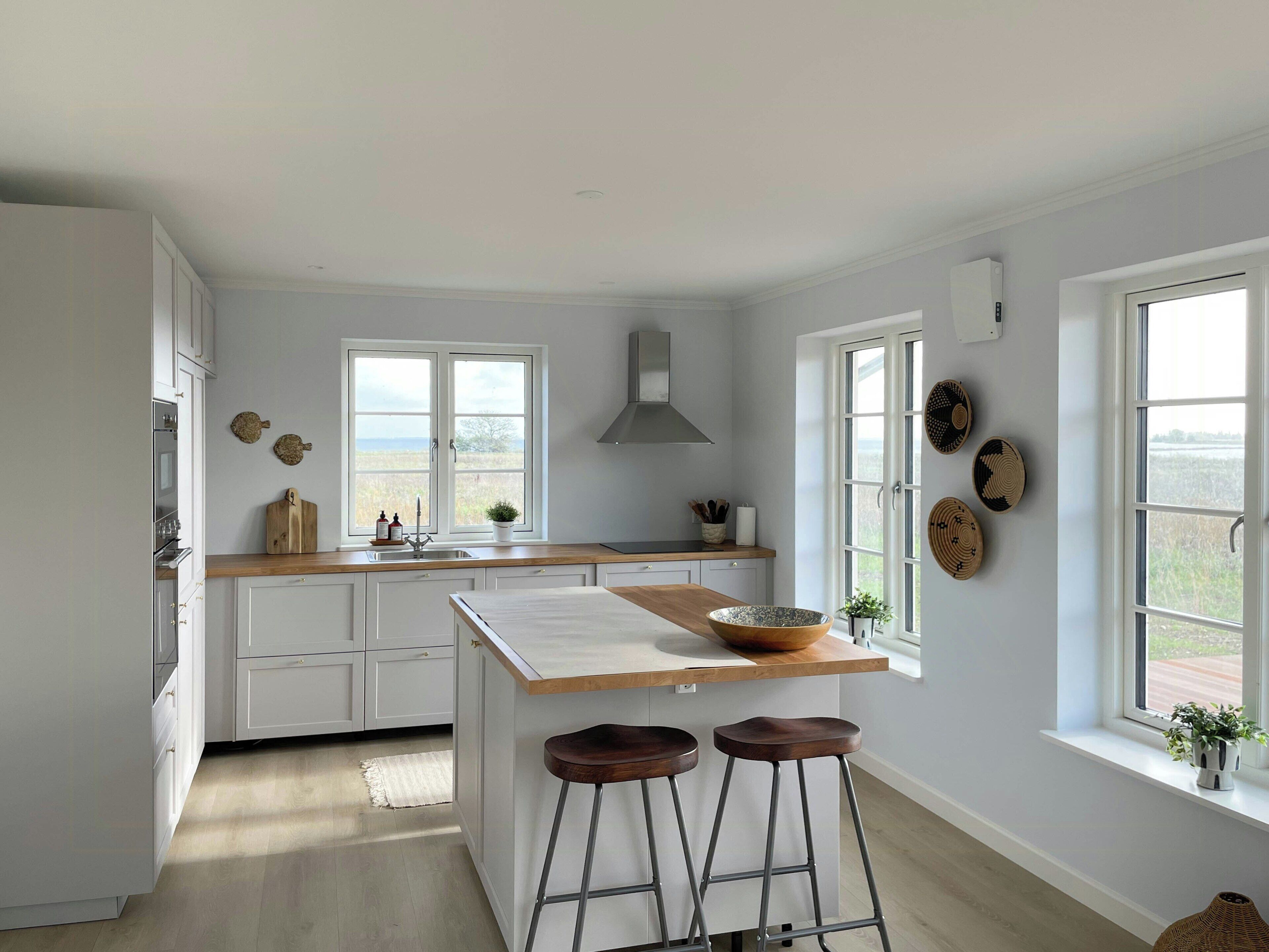 Une cuisine avec des meubles blancs, un plan de travail en bois, un îlot central avec des tabourets, une fenêtre offrant une belle vue et des assiettes décoratives accrochées au mur.
