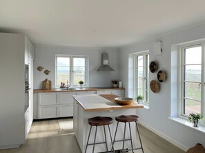 A kitchen with white cabinets, a wooden countertop, a central island with stools, a window with a view, and decorative plates on the wall. - House with great sea view on Lilleø (Lilleø)