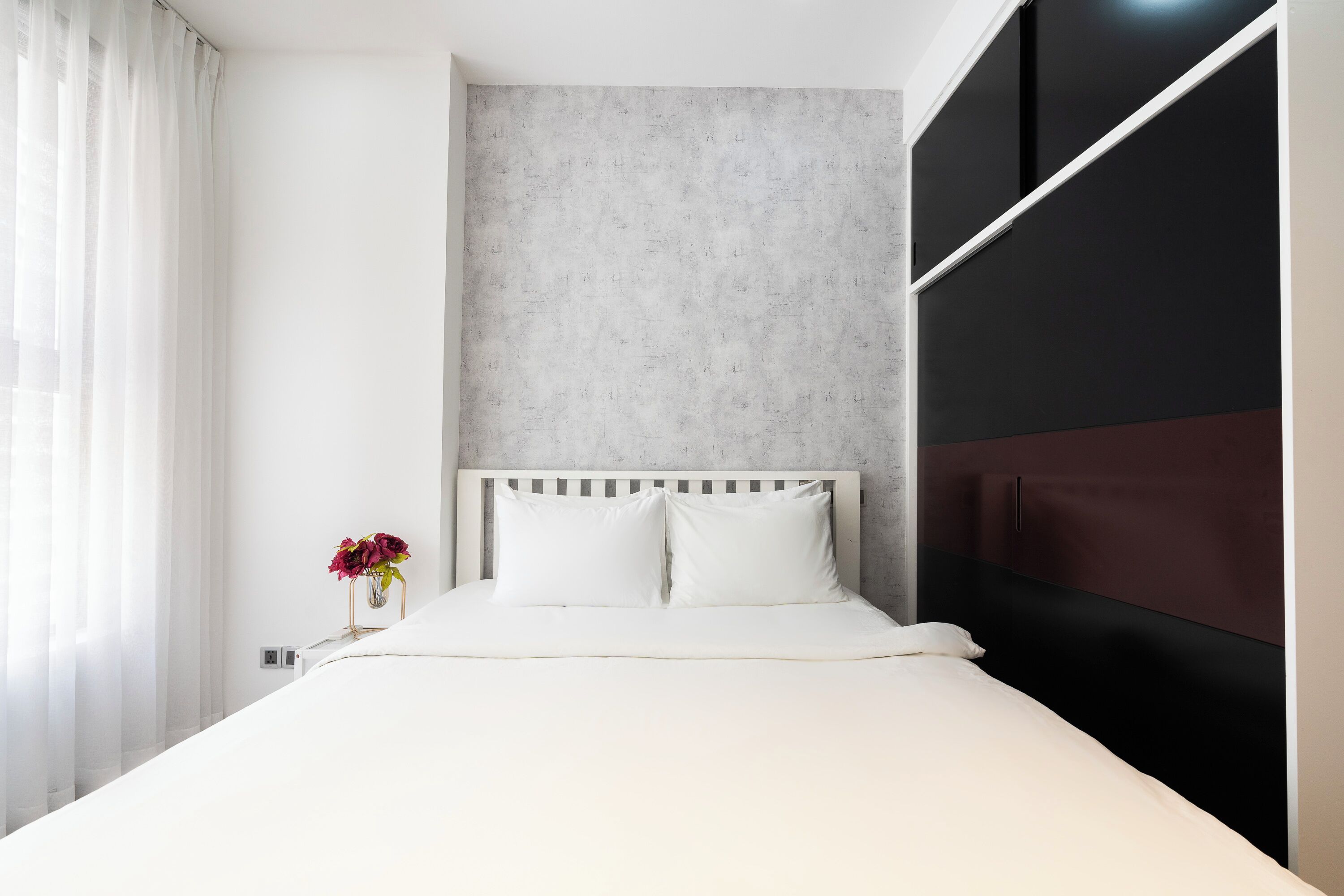 A modern hotel room with a bed, a nightstand, a vase with flowers, and a window with curtains.