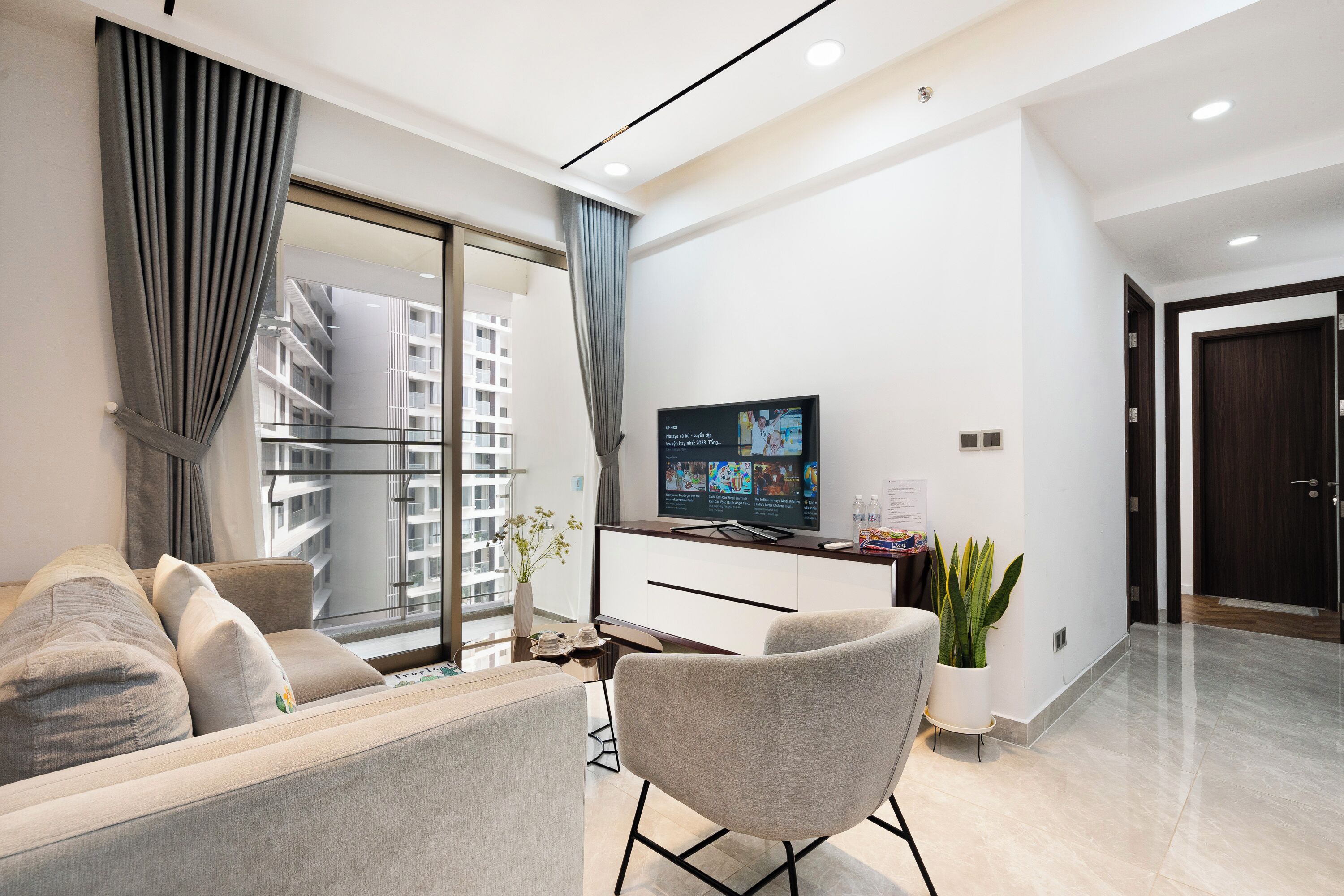 Een moderne woonkamer met een bank, een televisie, een plant en uitzicht op een gebouw door een groot raam.
