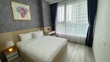 Une chambre d’hôtel moderne avec un grand lit, des tables de chevet, une petite table, une fenêtre avec des rideaux légers et une œuvre d’art florale encadrée accrochée au mur.