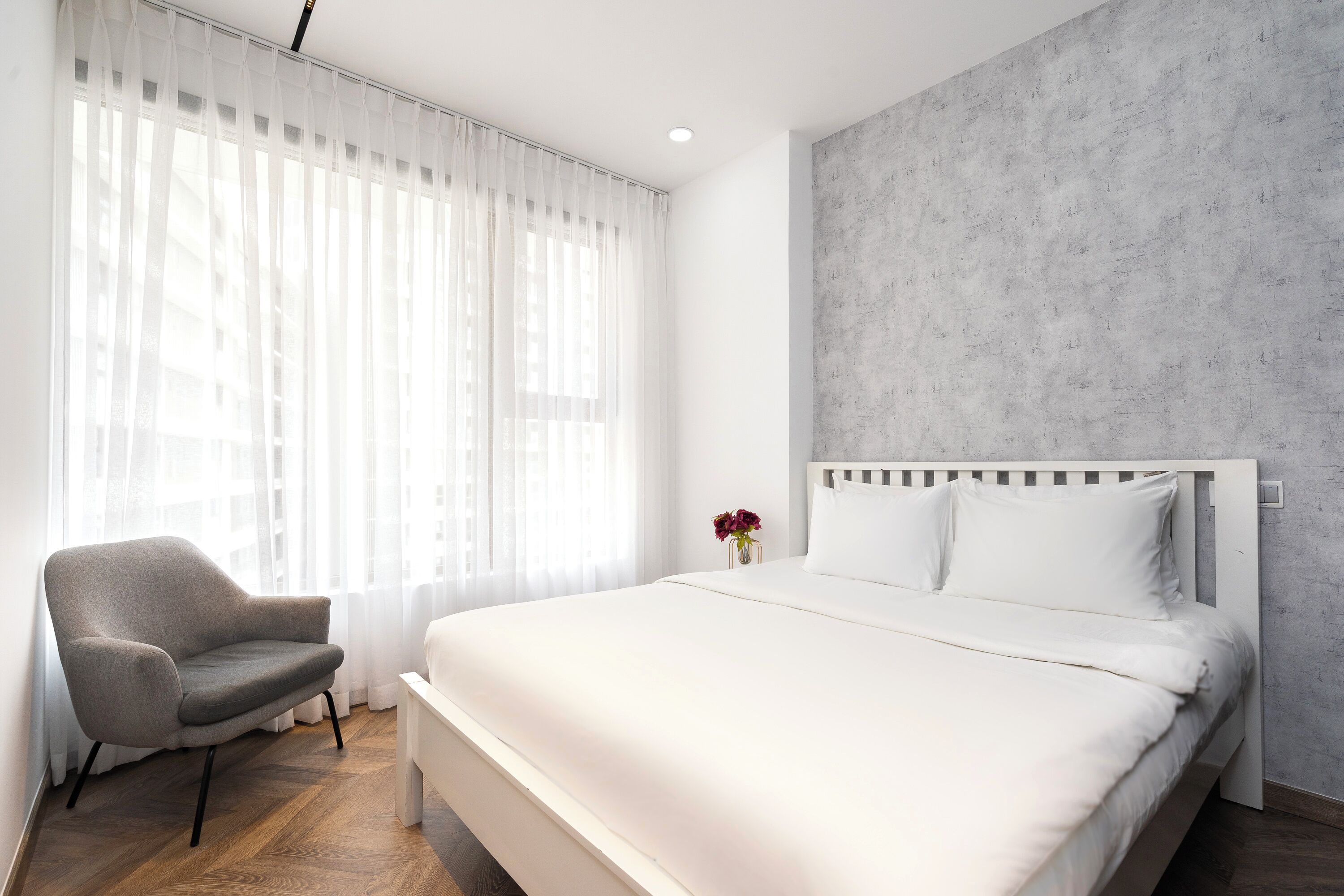 A modern bedroom with a large bed, a chair, and a window with sheer curtains.