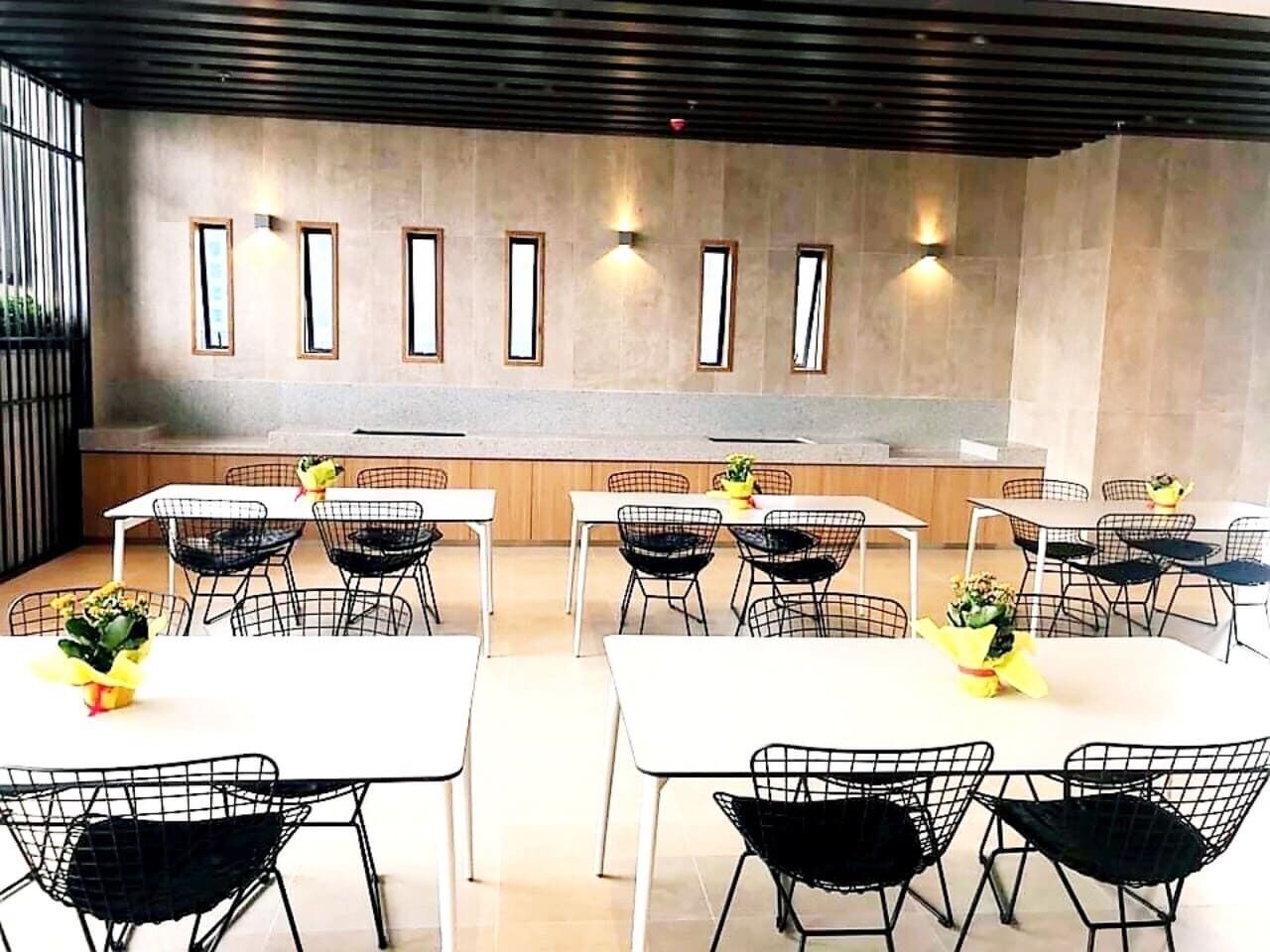 A modern dining area with white tables and black wire chairs, featuring a concrete wall with recessed lighting and a wooden ceiling.