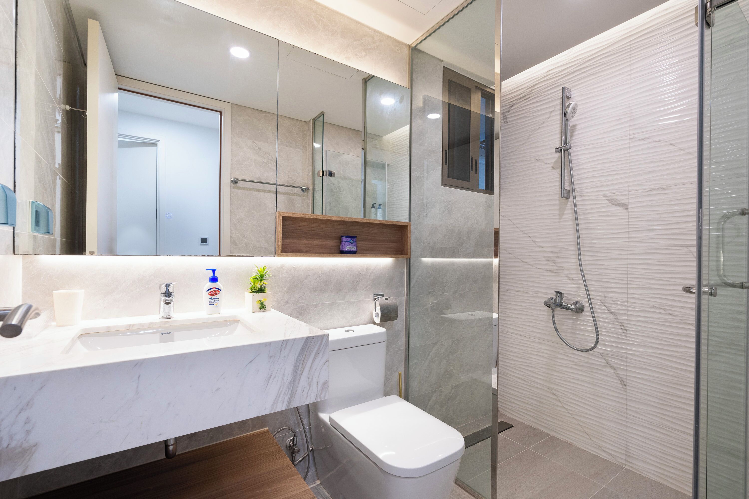 Un baño moderno con un lavabo de mármol, un inodoro, una cabina de ducha de vidrio y un estante de madera con artículos de aseo.