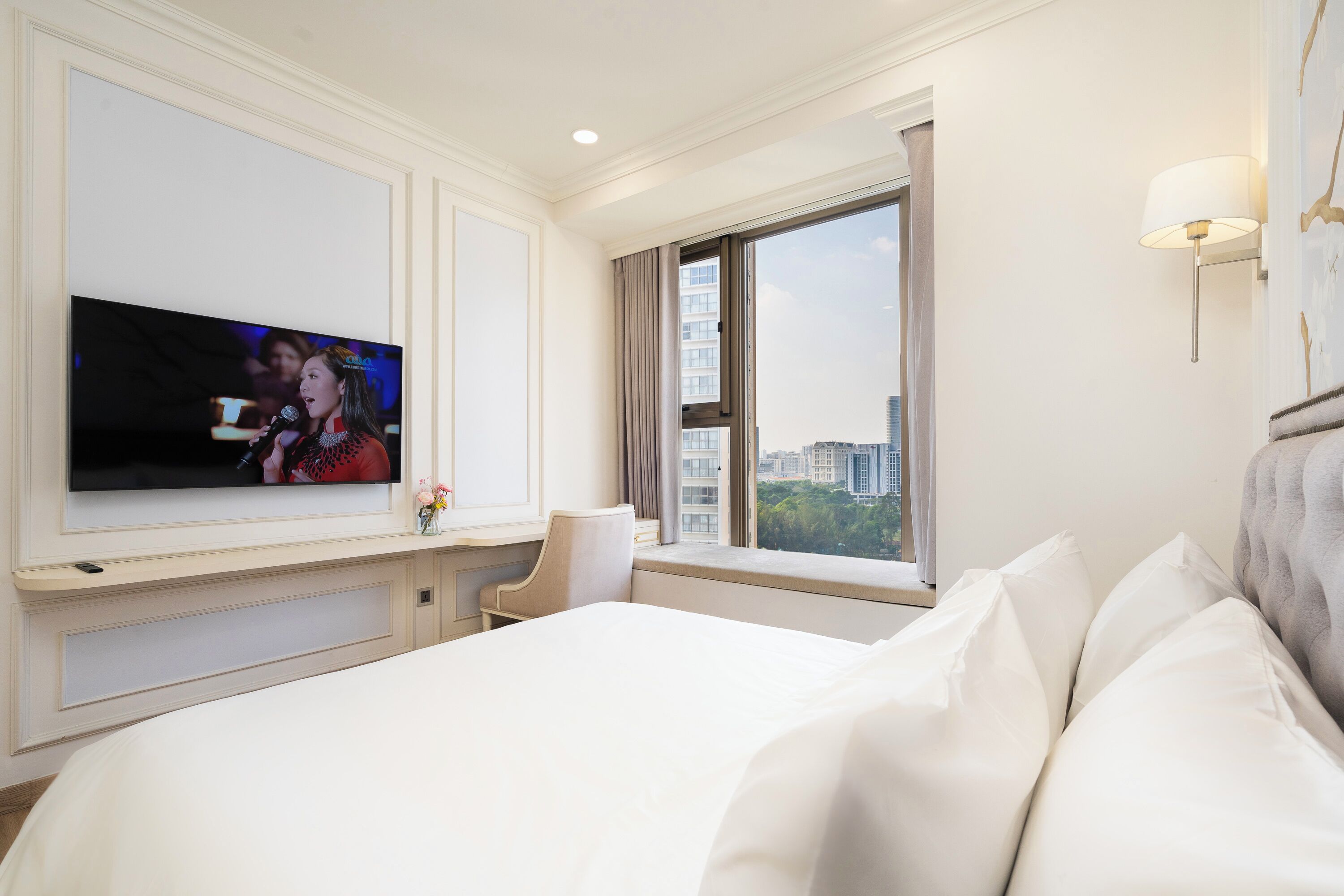 A hotel room with a large bed, a television on the wall, a window with a view of buildings, and a lamp on the wall.