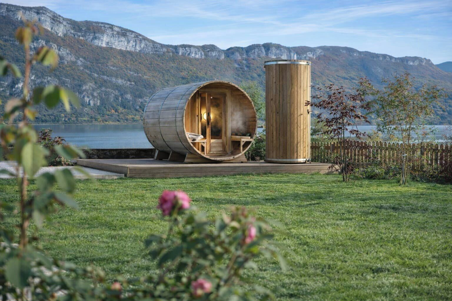 A cylindrical wooden structure with a bench inside, set on a wooden deck by a lake with mountains in the background.