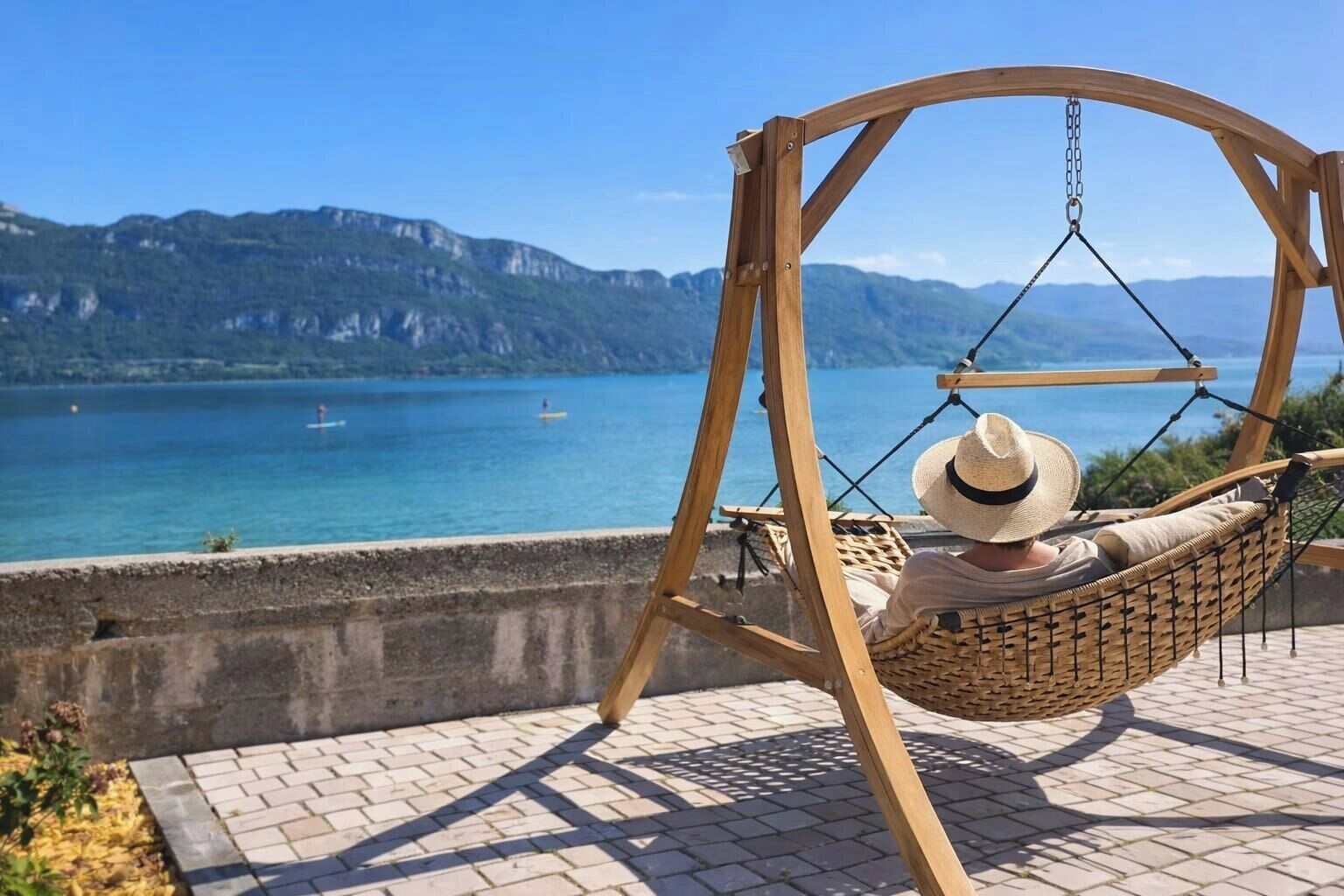 A wooden swing chair with a woven seat and backrest, positioned on a paved terrace overlooking a body of water with mountains in the background.