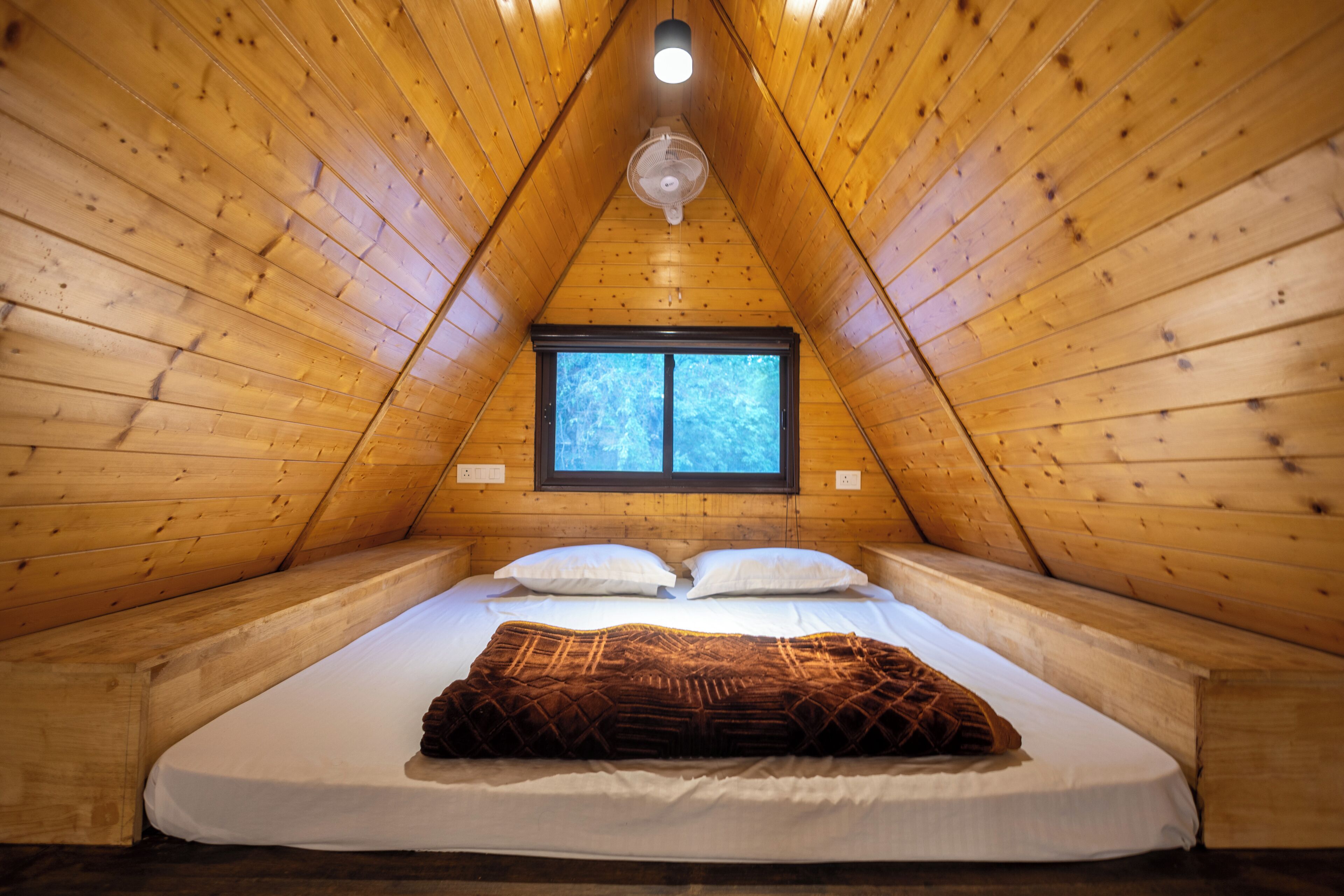 A room with a sloped ceiling, a bed with white bedding, two pillows, and a brown blanket. There is a window with a view of trees.