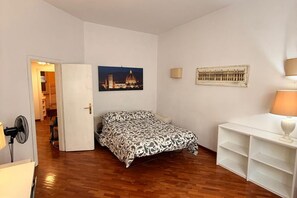 Een slaapkamer met een bed, een bureau, een stoel, een lamp, een foto aan de muur en een deur naar een andere kamer.