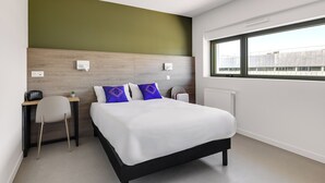A modern hotel room with a bed, a desk, a chair, a radiator, and a window.