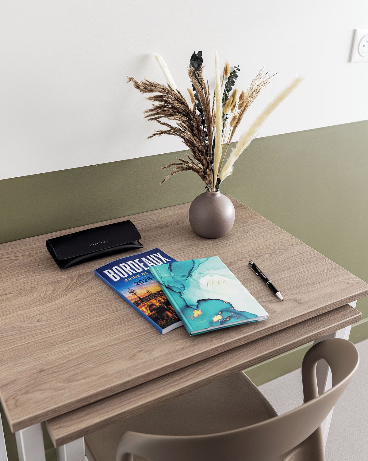 A wooden desk with a vase of dried plants, a black smartphone, a travel guidebook, and a pen.