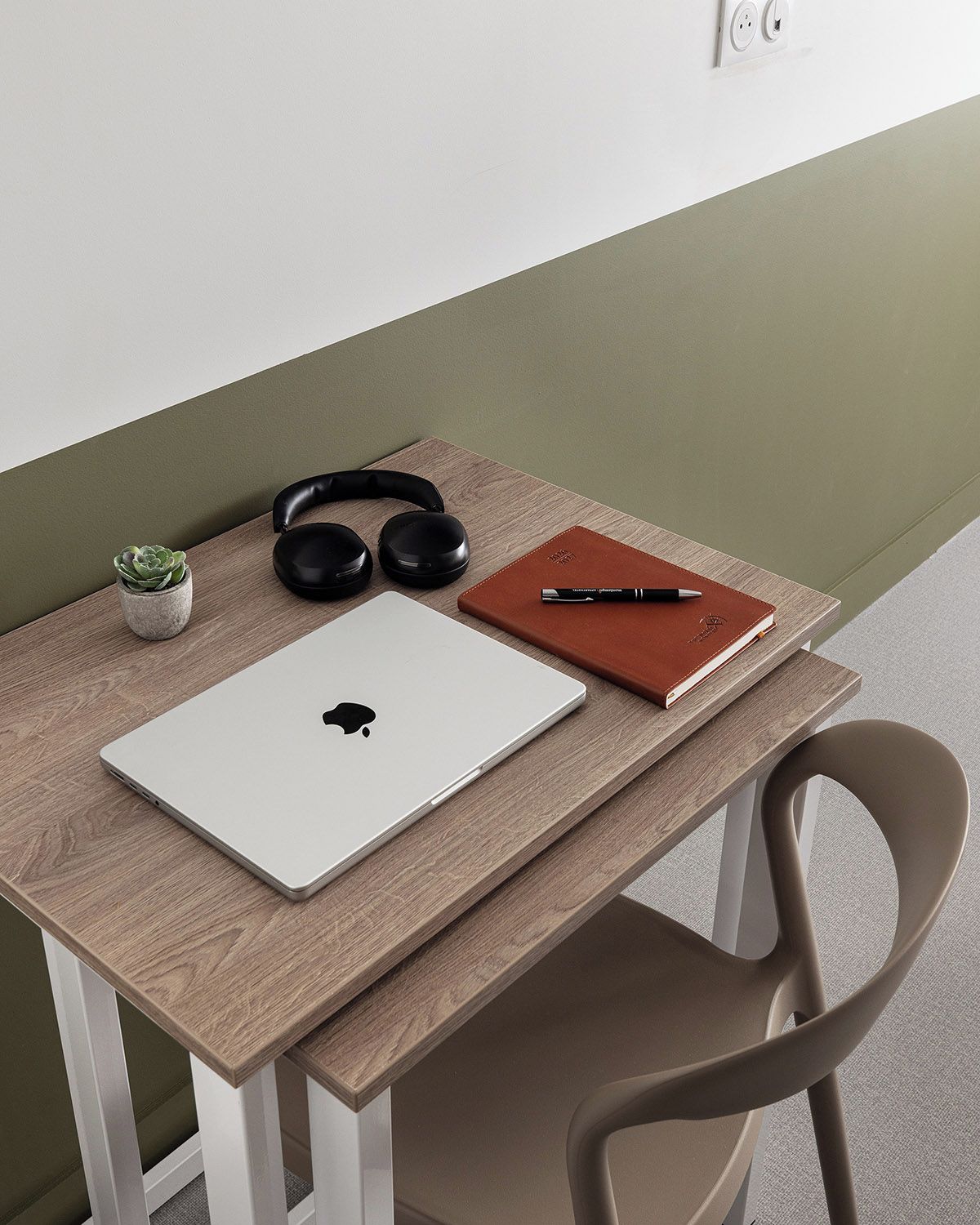 A wooden desk with a laptop, headphones, a notebook, and a pen.