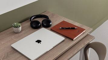 A wooden desk with a laptop, headphones, a notebook, and a pen.