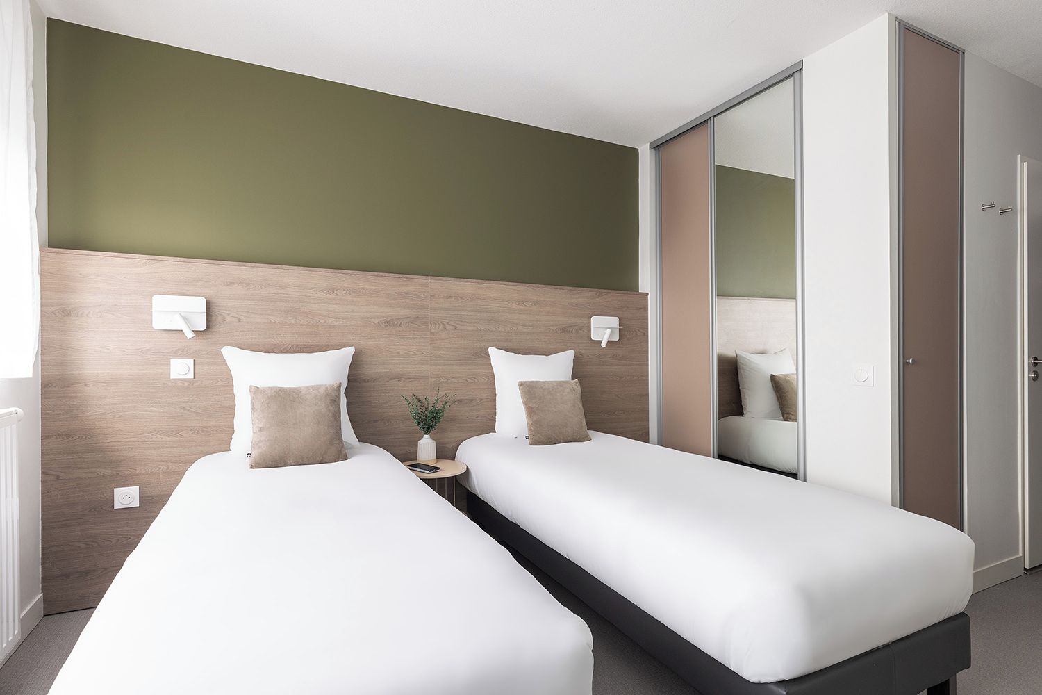 Ein Hotelzimmer mit zwei Einzelbetten, einem Nachttisch mit Pflanze, einem Spiegel und einem Kleiderschrank.