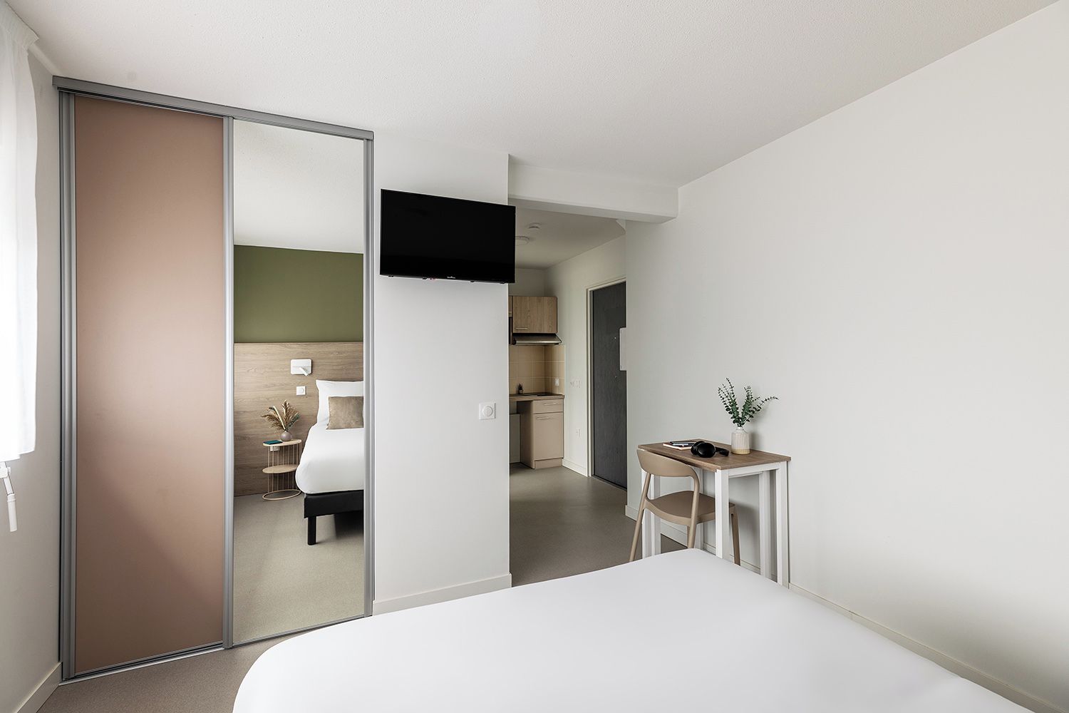 A modern hotel room with a large bed, a desk with a plant, and a view into the adjacent room.