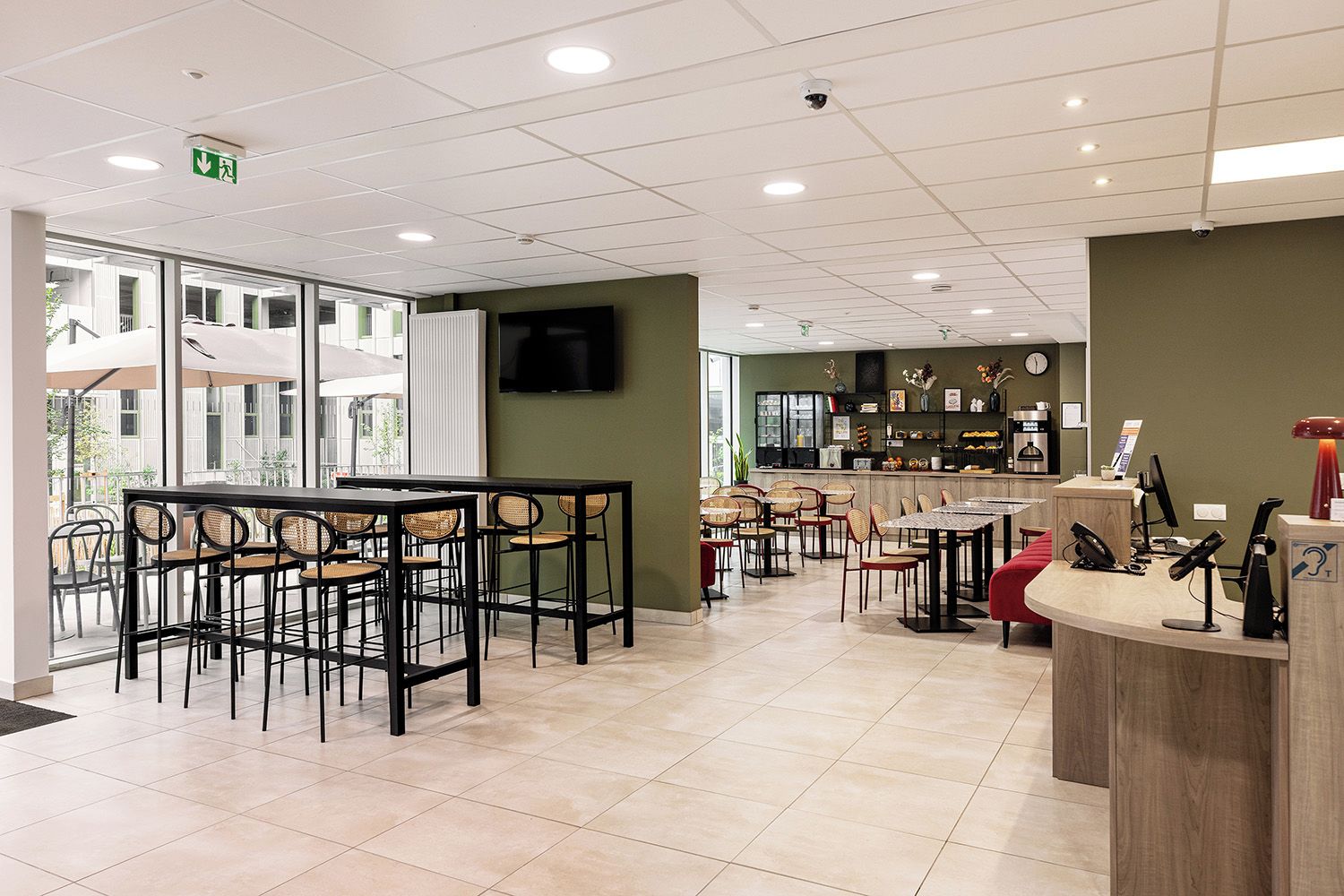 A cafe with multiple tables and chairs, a counter with a cash register, and a television mounted on the wall.