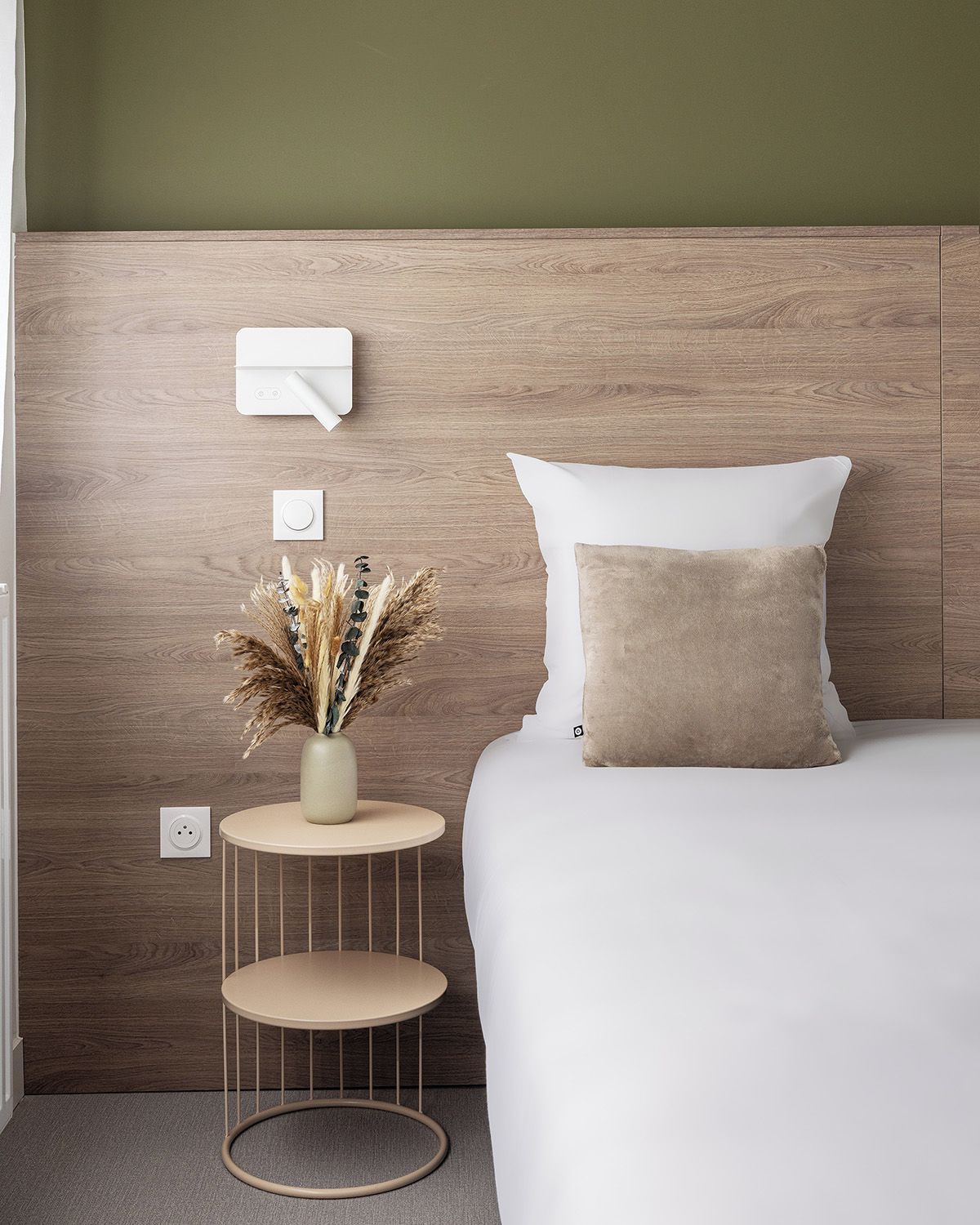 Une chambre à coucher avec une tête de lit en bois, une table de chevet ornée d’un vase contenant des plantes séchées, et un lit avec des oreillers blancs et beiges.