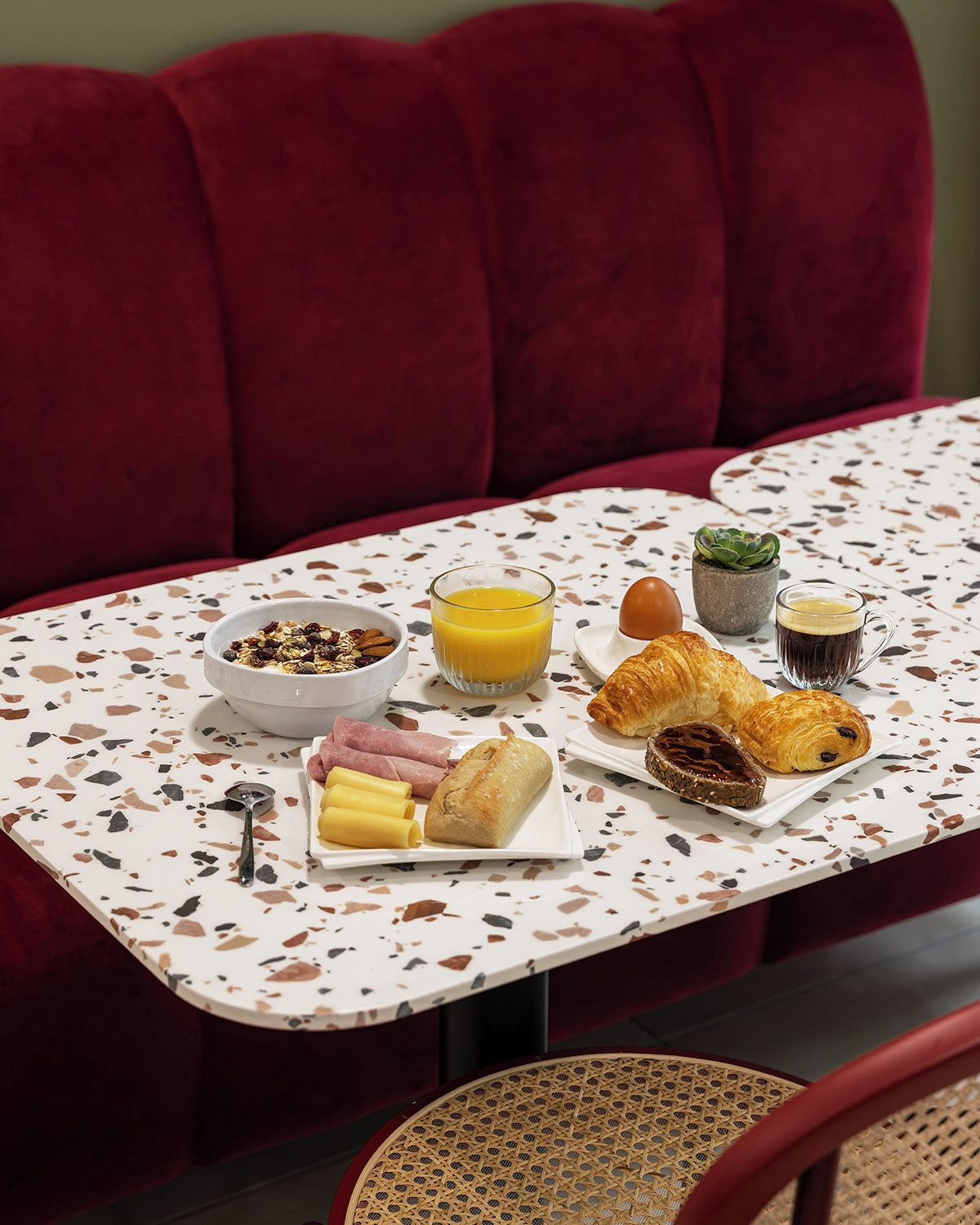 A breakfast setting with a table, red upholstered seating, and a variety of food items including pastries, a bowl of granola, and a glass of orange juice.