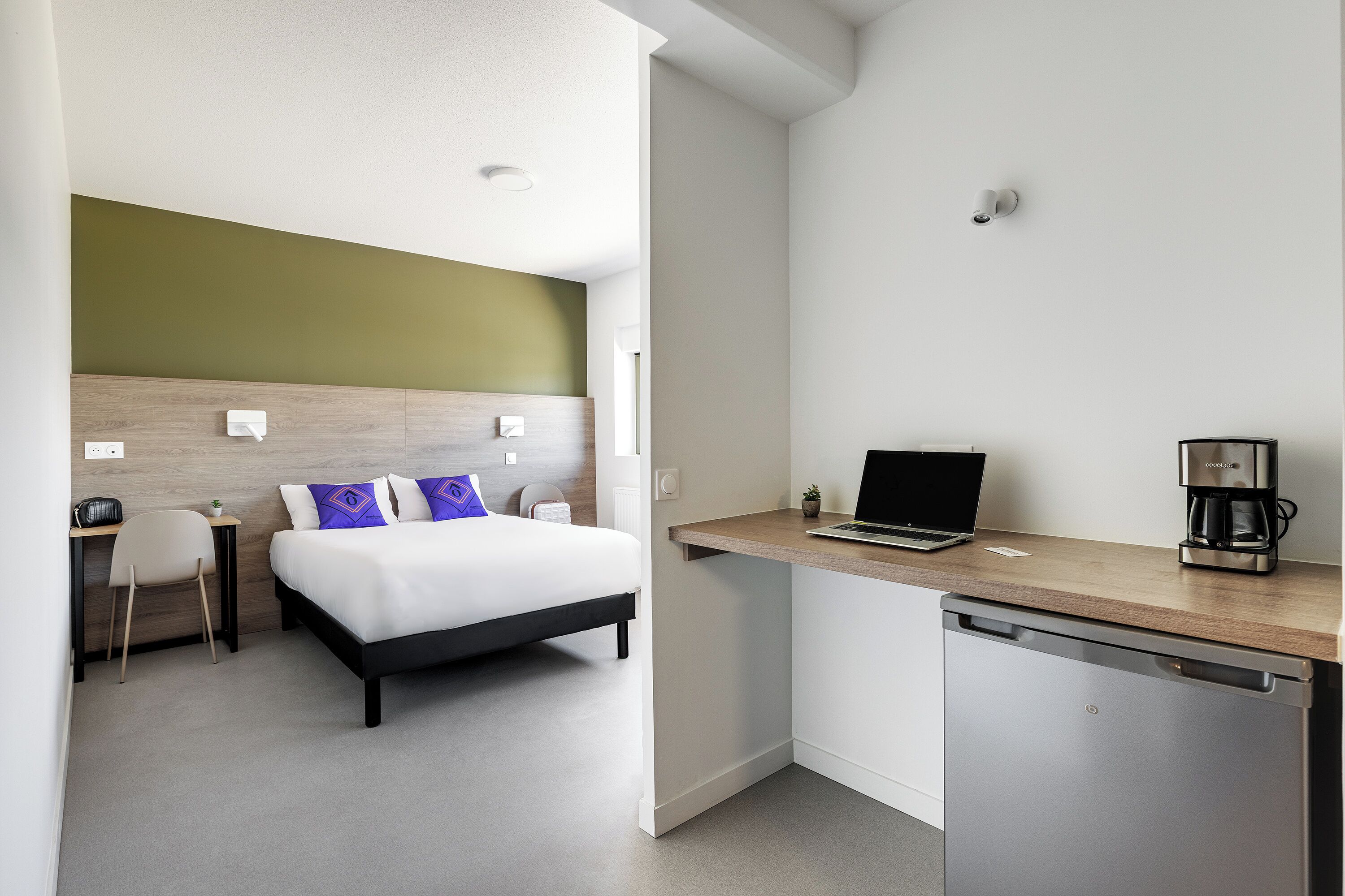 A compact hotel room with a bed, desk, chair, laptop, coffee maker, and dishwasher.