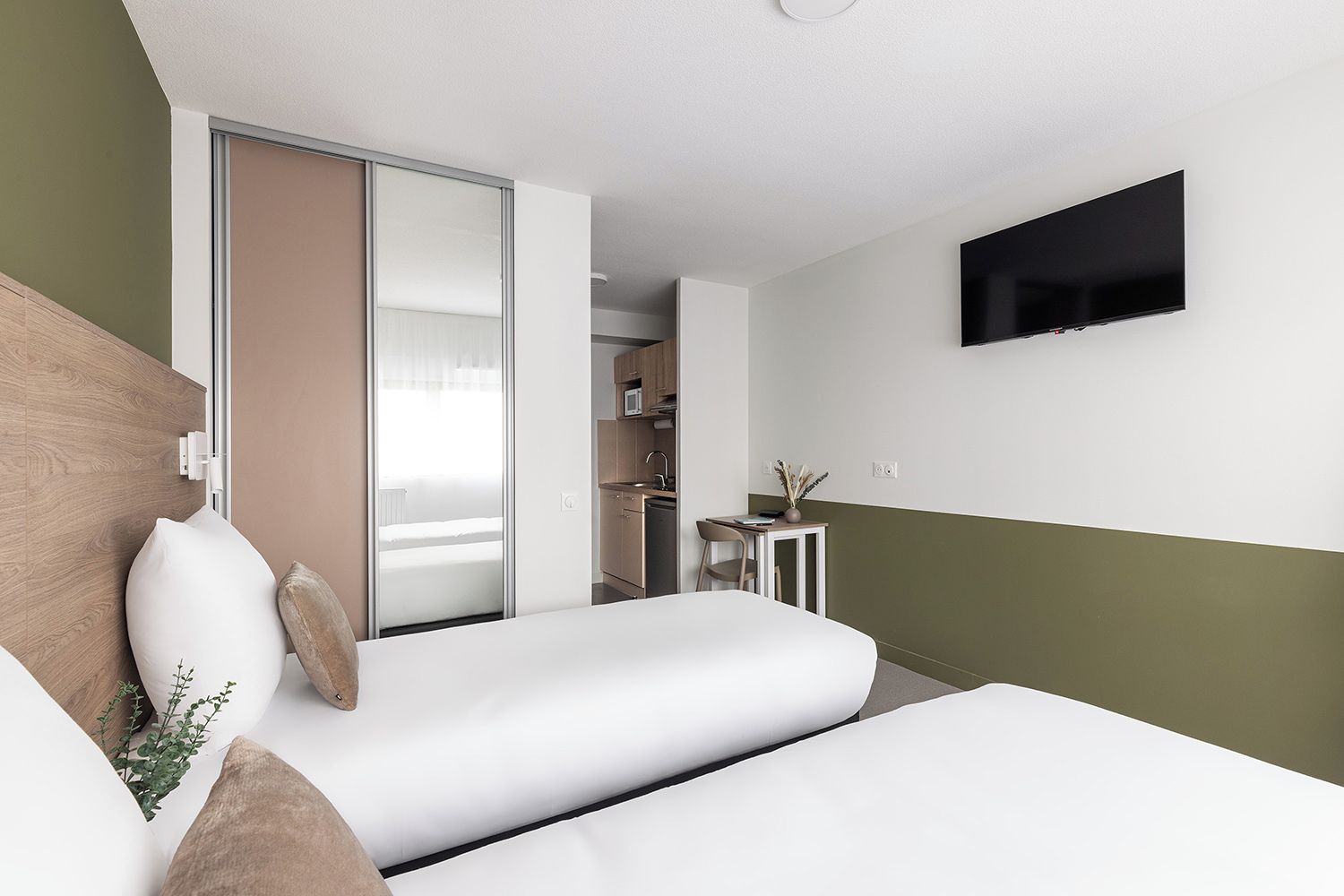 A modern hotel room with a bed, a TV mounted on the wall, and a kitchenette in the background.